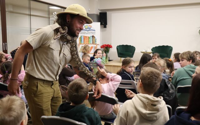 Na zdjęciu znajduje się mężczyzna wśród dzieci, którym daje dotknąć trzymanego przez siebie węża. W tle znajduje się mała scena z krzesłami oraz baner z logiem biblioteki i książkami.