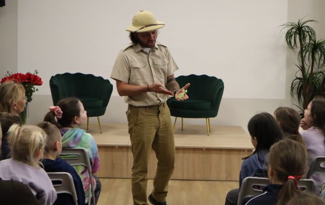 Na zdjęciu znajduje się mężczyzna, który ma na ręce liśćca, a drugą ręką na niego wskazuje, przed nim siedzą dzieci na krzesłach, a za nim znajduje się scena z zielonymi fotelami.