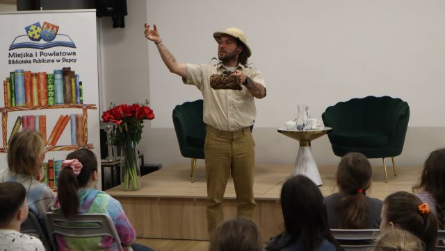 Na zdjęciu znajduje się mężczyzna trzymający w jednej ręce kore drzewa, a drugą rękę ma uniesioną do góry. Za mężczyzną znajdują się fotele i stolik, a przed nim grupa dzieci na krzesłach.