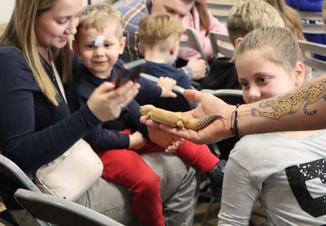 na zdjęciu dłoń, a na niej jaszczurka, wokół siedzące dzieci patrzące na zwierzę