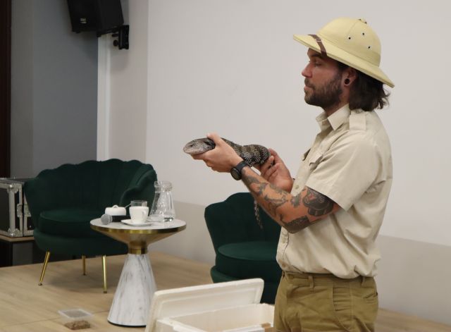 mężczyzna w stroju podróżnika trzymający w rękach dużą jaszczurkę, w tle na scenie zielone fotele i stolik