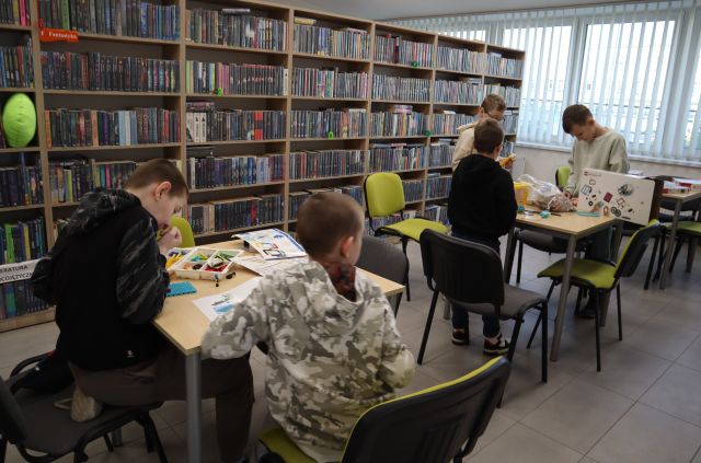 Na zdjęciu znajduje sie piatka chłopców, dwóch przy pierwszym stoliku i trzech przy drugim. Chłopcy budują konstrukcje z klocków LEGO. W tle znajdują się półki z książkami.