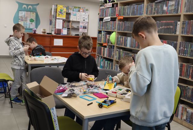 Na zdjęciu znajduje się trzech chłopców przy stoliku, dwóch z nich stoi i układają LEGO, za nimi znajduje się dójka chłopców przy stoliku, którzy także tworzą swoje konstrukcje z LEGO. W tle znajdują się półki z książkami i biurko.