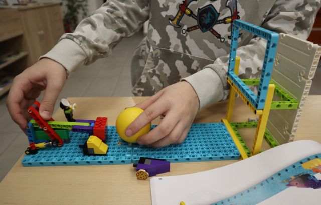 Na zdjęciu znajduje się konstrukcja z LEGO wraz z żółtą piłeczką, na której rękę ma chłopiec poprawiający swoją konstrukcje.