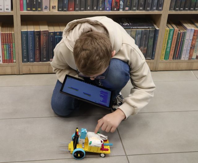 Na zdjęciu znajduje się chłopiec klękający na podłodze z tabletem w jednej ręce, drugą dotyka swojego robota z LEGO. W tle znajdują się półki z książkami.