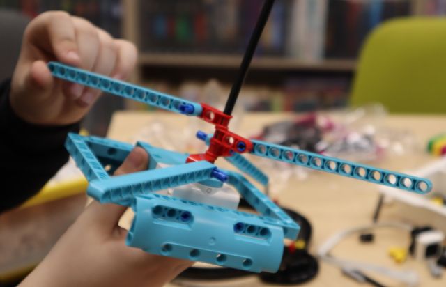 Na zdjęciu znajduje się czubek helikoptera zbudowanego z klocków LEGO, konstrukcja jezt trzymana przez uczestnika zajęć, na zdjęciu uchwycone zostały tylko ręce. W tle znajduje się stół, na którym leżą klocki, kawałek krzesła oraz półki z książkami.