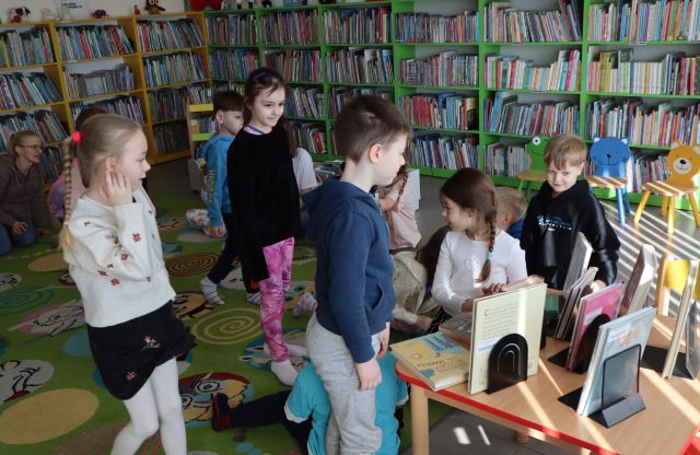 Na zdjęciu znajdują się dzieci, część stoi, część klęczy przed stolikiem, na którym wystawione są książkę. Dzieci znajdują się na dywanie, a dookoła znajdują się półki z książkami.
