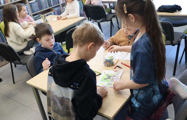 Na zdjęciu w centrum znajduje się czwórka dzieci kolorująca przy stoliku. Dookoła znajduje się reszta przedszkolaków przy stolikach oraz półki z książkami w tle.