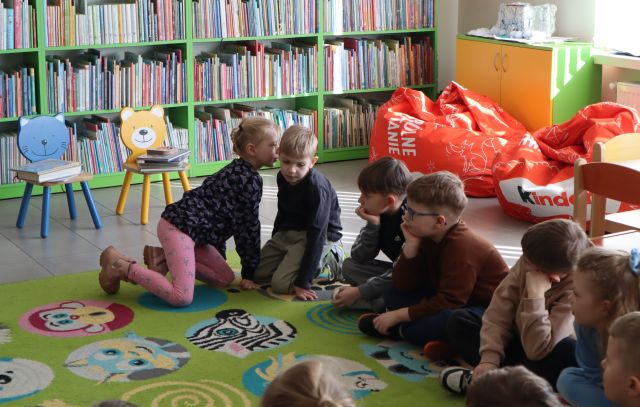Na zdjęciu znajdują się siedzące na dywanie obok siebie dzieci, dziewczynka mówi coś chłopcu obok na ucho. W tle znajdują się półki z książkami i pufy.