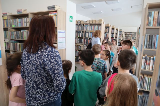 Na zdjęciu znajdują się dzieci w bibliotece między półkami, na których znajdują się książki. Po lewej stronie stoi kobieta tyłem.