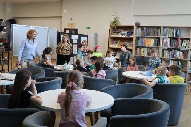Na zdjęciu znajdują się dzieci w czytelni, część z nich siedzi przy stolikach na fotelach. W oddali znajdują się dwie kobiety, półki na gazety i książki.