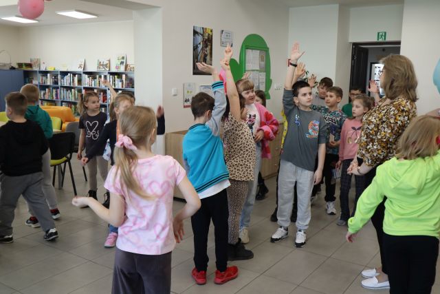 Na zdjęciu znajdują się dzieci, część z nich ma podniesioną rękę. Stoją oni na wejściu do sali, obok nich znajduje się przejście do pomieszczenia z książkami. Pomiędzy nimi po prawej stronie stoi kobieta.