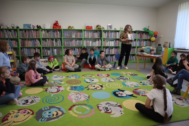 Na zdjęciu znajdują się dzieci część z nich siedzi na dywanie, część na pufach przy nim. Kobieta stojąca między dziećmi trzyma w rękach książkę i stoi obok stoliczka, na którym są poukładane książki. W tle znajdują się półki z książkami.
