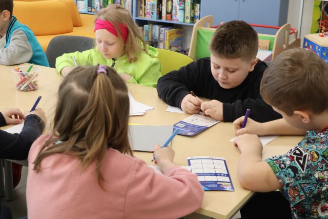 Na zdjęciu znajduje się czwórka drugoklasistów przy złączonych stolikach i piszą na kartkach. W tle znajdują się półki z grami, kawałek źółtej kanapy.