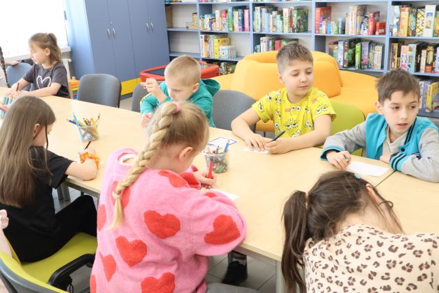 Na zdjęciu znajdują się dzieci przy złączonych stolikach i piszą na kartkach. W tle znajdują się półki z książkami i żółta okrągła kanapa.