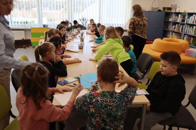 Na zdjęciu znajdują się dzieci siedzące przy złączonych stolikach i rysują na kartkach. Po dwóch stronach stołu stoją kobiety. W tle znajdują się okna.