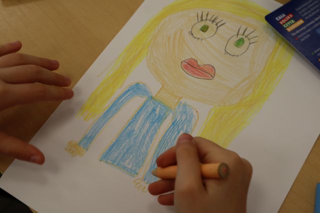 Na zdjęciu znajduję się ilustracja dziewczyny o blond włosach. Przy obrazku znajdują się dwie dziecięce ręce.