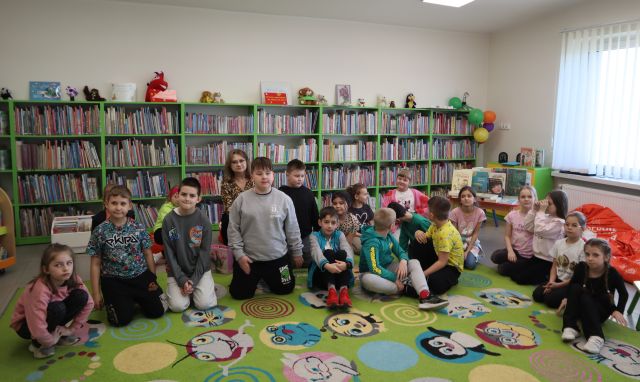 Na zdjęciu znajduje się dziewiętnaście dzieci oraz jedna kobieta, siedzących na dywanie do wspólnego zdjęcia. W tle znajdują się półki z książkami.