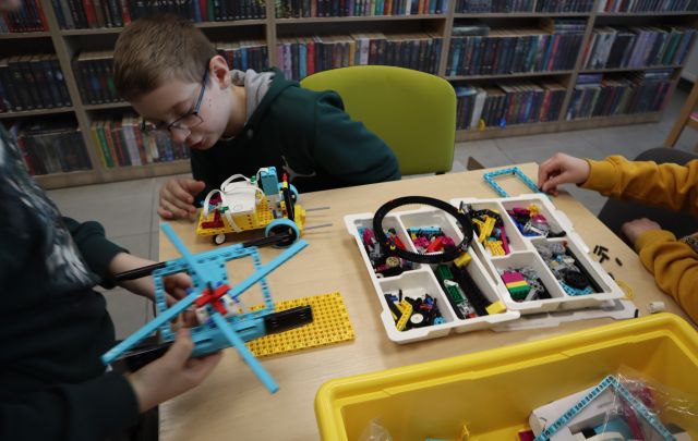 Na zdjęciu znajduje się stolik, na którym leżą klocki LEGO oraz konstrukcje budowane przez chłopców siedzących przy stoliku. W tle znajdują się półki z książkami.