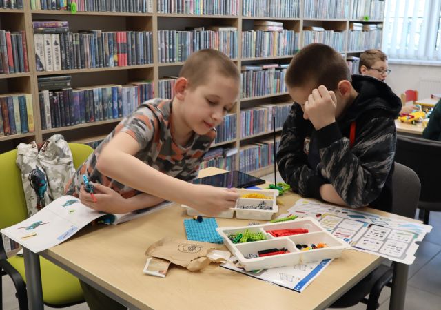 Na zdjęciu znajduje się dwójka chłopców, którzy wspólnie układają LEGO przy stoliku. W tle znajdują się półki z książkami.
