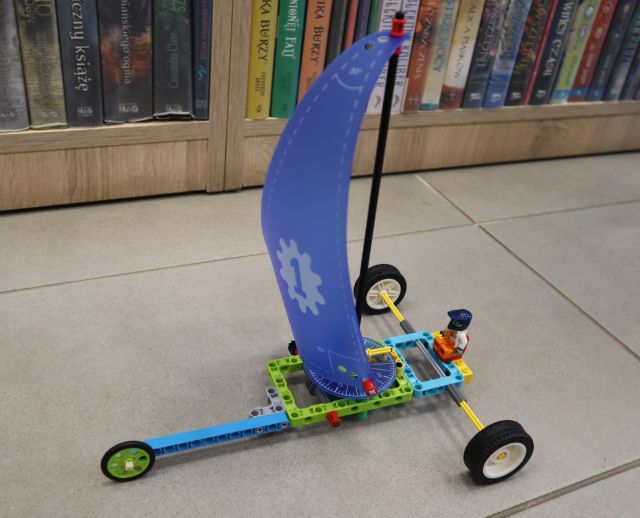 Na zdjęciu znajduje się zbudowany z LEGO, posiada niebieski żagiel. Konstrukcja leży na podłodze, a w tle znajdują się półki z książkami.