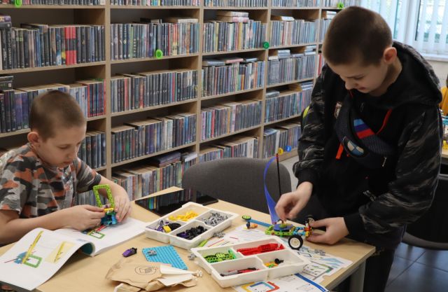 Na zdjęciu znajduje się dwójka chłopców przy stoliku, jeden siedzi drugi stoi i wspólnie układają LEGO. W tle znajdują się półki z książkami.