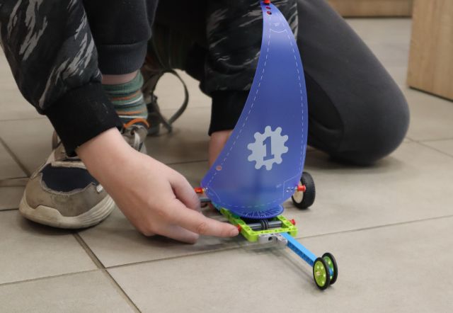 Na zdjęciu znajduje się chłopiec, któzy trzyma znajdujący się na podłodze pojazd z LEGO z żaglem.