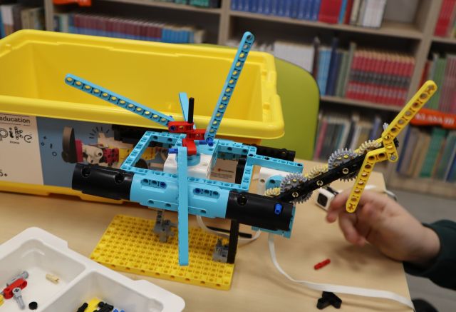 Na zdjęciu znajduje się zbudowany z LEGO helikopter leżący na stoliku, za nim znajduje się żółte pudełko, a po prawej stronie przy stoliku znajduje się dziecięca ręka.
