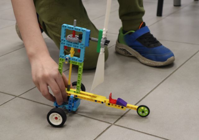 Na zdjęciu znajduje się konstrukcja z LEGO na podłodze trzymana przez klęczącego chłopca.