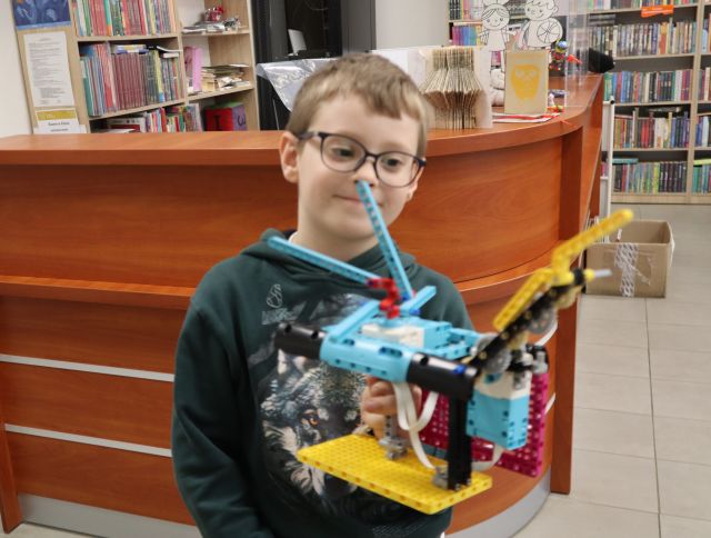 Na zdjęciu znajduje się chłopiec trzymający w ręce zbudowany przez siebie helikopter z klocków LEGO, za nim znajduje się biurko o raz półki z książkami.