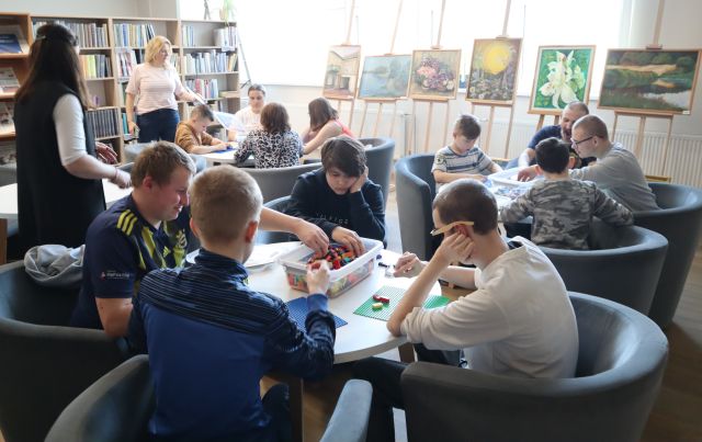 Na zdjęciu znajduje się młodzież siedząca przy okrągłych stołach, przy których układają klocki LEGO. W tle znajdują się obrazy oraz okna, a po prawej stronie półki z książkami.
