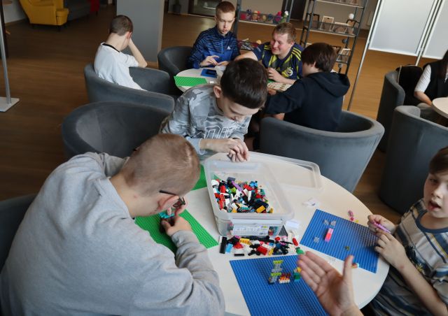 Na zdjęciu znajduje się młodzież układająca klocki LEGO przy okrągłych stolikach.