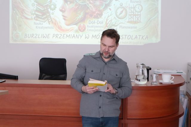 Na zdjęciu znajduje się mężczyzna z książką w ręku, który opiera się o biurko, w tle znajduje się wyświetlana na ścianie prezentacja.