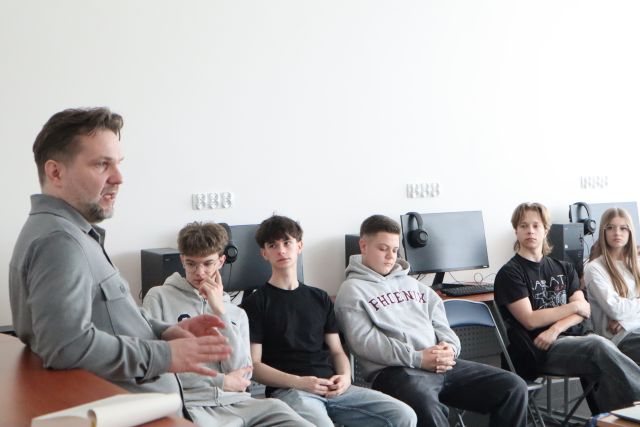 Na zdjęciu znajduje się mężczyzna bokiem, a obok znajduje się siedząca na krzesłach młodzież. Warsztaty odbywają się w sali komputerowej.