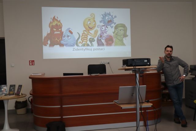 Na zdjęciu znajduje się mężczyzna opierający się o biurko po prawej stronie, na środku znajduje się rzutnik z laptopem i wyświetla na śniacie prezentacje przedstawiającą emocje.