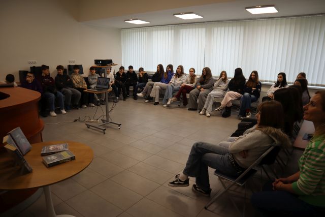 Na zdjęciu znajduje się młodzież siedząca na krzesłach ustawionych w literę U, na środku stoi rzutnik, a w tle znajdują się okna. Warsztaty odbywały się w sali komputerowej.