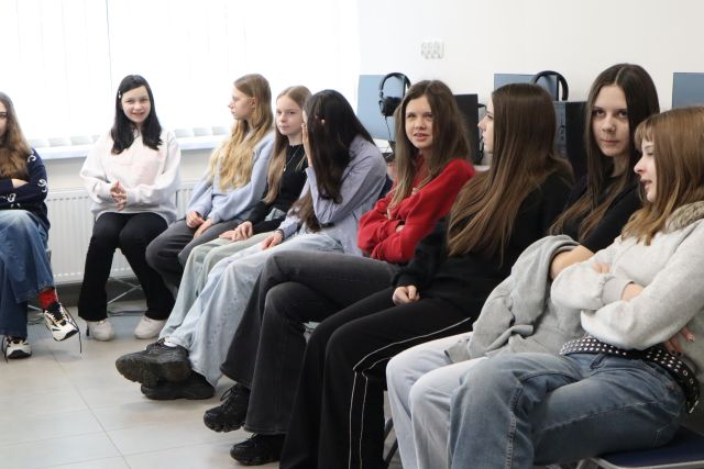 Na zdjęciu znajduje się młodzież siedząca na krzesłach w sali komputerowej.