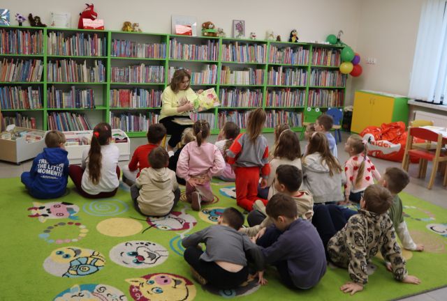 Na zdjęciu znajdują się siedzące na dywanie przedszkolaki, a przed nimi siedzi kobieta z książką. W tle znajdują się półki z książkami.
