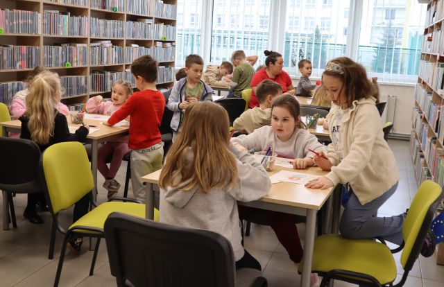 Na zdjęciu znajdują się przedszkolaki siedzące przy pięciu stolikach, między nimi znajdują się dwie kobiety. W tle znajdują się okna oraz półki z książkami po lewej stronie.