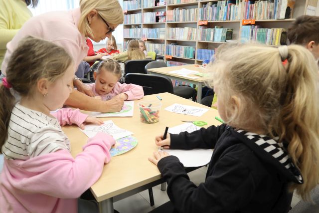 Na zdjęciu znajdują się trzy dziewczynki przy stoliku, przy jednej nachyla się kobieta i pomaga jej w kolorowaniu. W tle znajduje się reszta stołów oraz półki z książkami.
