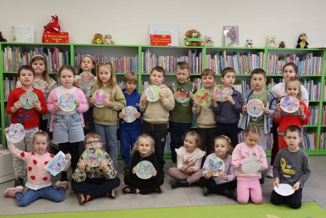Na zdjęciu znajduje się grupa przedszkolaków ze swoimi pracami, które przedstawiają motyle na łące. W tle znajdują się półki z książkami.
