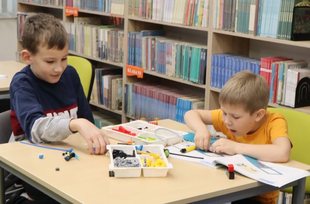 Na zdjęciu znajduje się dwójka chłopców przy stoliku, którzy wspólnie układają znajdujące się na stoliku klocki LEGO. W tle znajdują się półki z książkami.