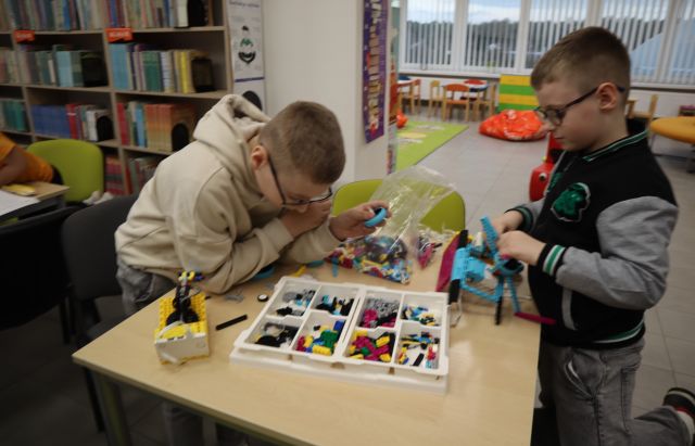 Na zdjęciu znajduje się dwójka chłopców stojących przy stoliku i wspólnie układają leżące na stole klocki LEGO. W tle znajdują się półki z książkami oraz przejście do kącika malucha.