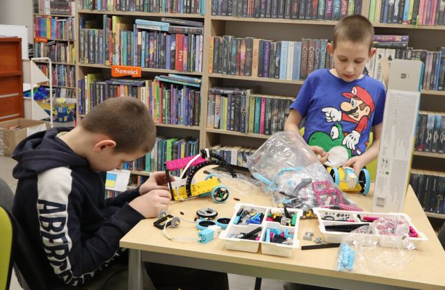 Na zdjęciu znajduje się dwójka chłopców, jeden siedzi drugi stoi. Na stoliku znajdują się klocki LEGO oraz karton, a w tle półki z książkami.