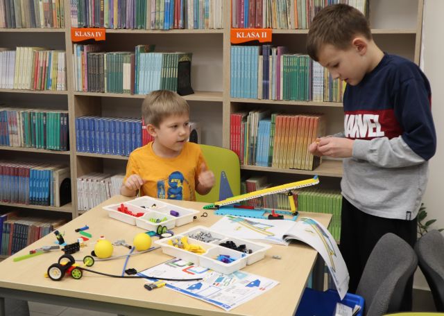 Na zdjęciu znajduje się dwójka chłopców przy stoliku jeden z nich stoi, na stole znajduje się zbudowana przez nich z klocków LEGO skocznia narciarska. W tle znajdują się półki z książkami.
