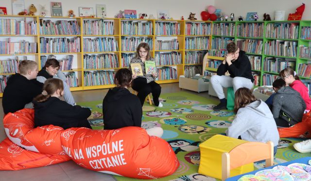 Na zdjęciu znajduje się młodzież siedząca na pufach na dywanie wraz z kobietą, która pokazuje im książkę. W tle znajdują się półki z książkami.