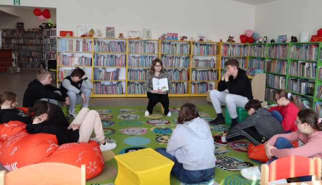 Na zdjęciu znajduje się kobieta wraz z dziewiątką nastolatków na dywanie. W tle znajdują się półki z książkami.