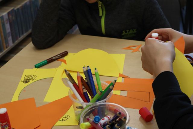 Na zdjęciu znajduje się stolik z żółtymi i pomarańczowymi kartkami oraz kredkami, po prawej stronie widoczne są dłonie trzymające żółtą kartkę.