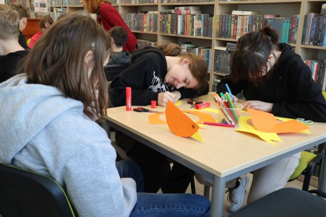 Na zdjęciu znajduje się trójka dziewczyn przy stoliku, na którym leżą kolorowe kartki, kredki i wykonne przez nie kurczaki z papieru. W tle znajdują się półki z książkami.