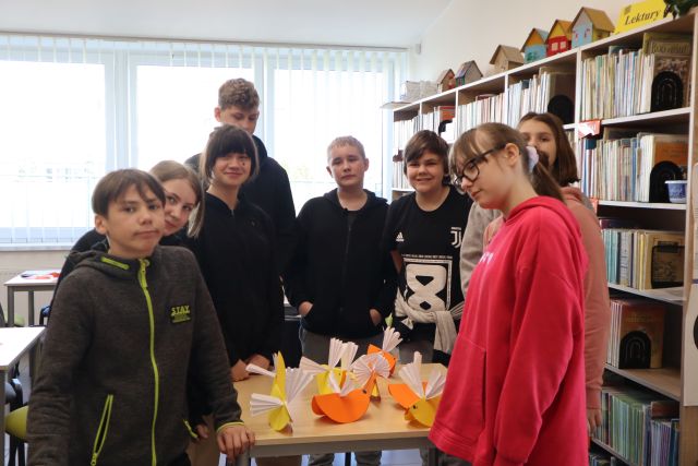 Na zdjęciu znajduje się dziewiątka młodzieży wśród stolika, na którym leżą wykonane przez nich kurczaki. W tle znajdują się okna oraz półki z książkami.